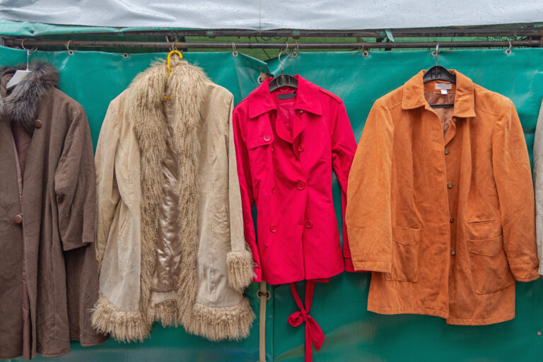 Im Hintergrund hängt eine türkisfarbene Plane an einer Kleiderstange. Davor hängen vier Kleidungsstücke nebeneinander an Kleiderbügeln: Ein brauner Mantel mit hoffentlich Kunst-Fell-Kragen, ein beiger Hippie-Mantel mit Fransen, ein knallroter Trenchcoat und eine orangefarbenes Cordhemd.