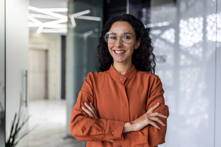 Young beautiful and successful hispanic businesswoman smiling and looking at camera, female worker working inside office wearing glasses and curly hair, arms crossed portrait